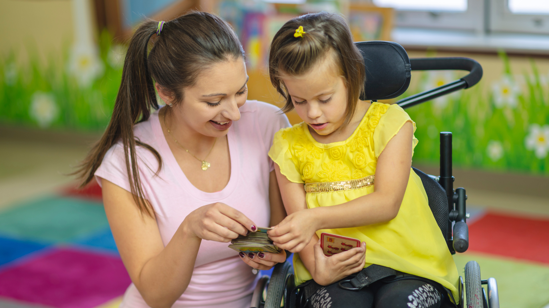 A young woman and a girl using early mobility supports play a card game together in a colorful classroom. Both are smiling and focused on the cards, sharing a joyful and engaging moment while moving to learn.