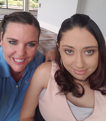 Two women taking a selfie indoors; one is smiling in a blue shirt, and the other, featured in Angelas Story, has long dark hair, hoop earrings, and wears a light pink top while looking softly at the camera.