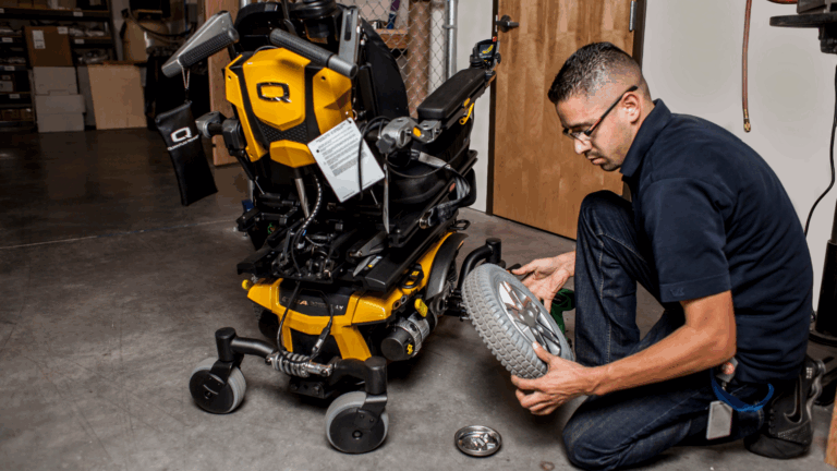 A man kneels on the floor, inspecting the wheel configuration of a yellow and black powered wheelchair in a workshop. Tools and parts are visible nearby.