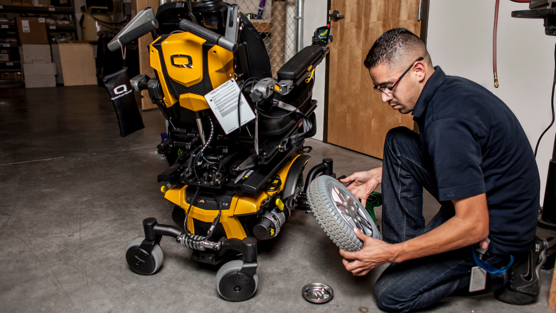 A man kneels on the floor, inspecting the wheel configuration of a yellow and black powered wheelchair in a workshop. Tools and parts are visible nearby.