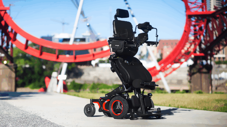 A modern black and red electric wheelchair with a headrest and elevated seat is positioned on a paved path outdoors, symbolizing effective advocacy, with a large red bridge structure and buildings visible in the background.