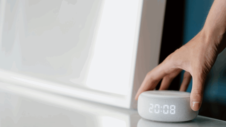 A hand adjusts a small, round smart speaker with a digital clock display reading 20:08, placed on a white surface next to a bright rectangular object, showcasing smart home technology.