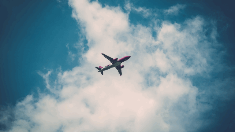 A commercial airplane with a pink and white color scheme flies overhead against a partly cloudy blue sky, symbolizing the freedom and possibilities of air travel for everyone, including those using a power wheelchair.