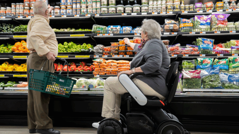 An older man holds a shopping basket while standing beside a woman in a wheelchair as she reaches for vegetables in the grocery store produce aisle, highlighting the importance of accessible grocery shopping.
