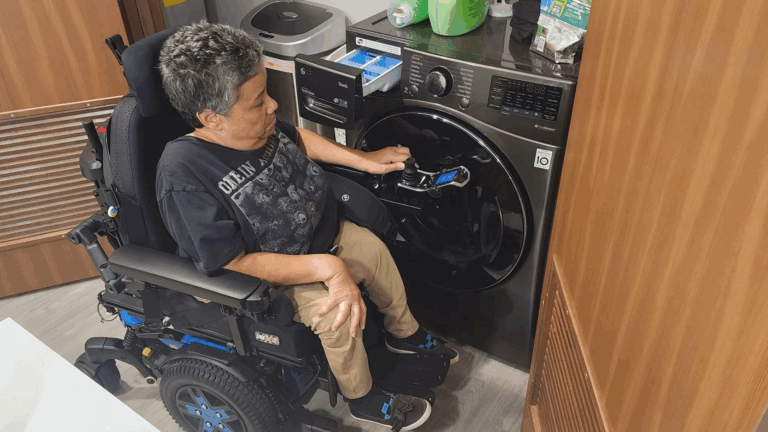 A person in a motorized wheelchair loads laundry into a front-loading washing machine in a compact, accessible room with wooden cabinets and cleaning supplies on top of the washer.