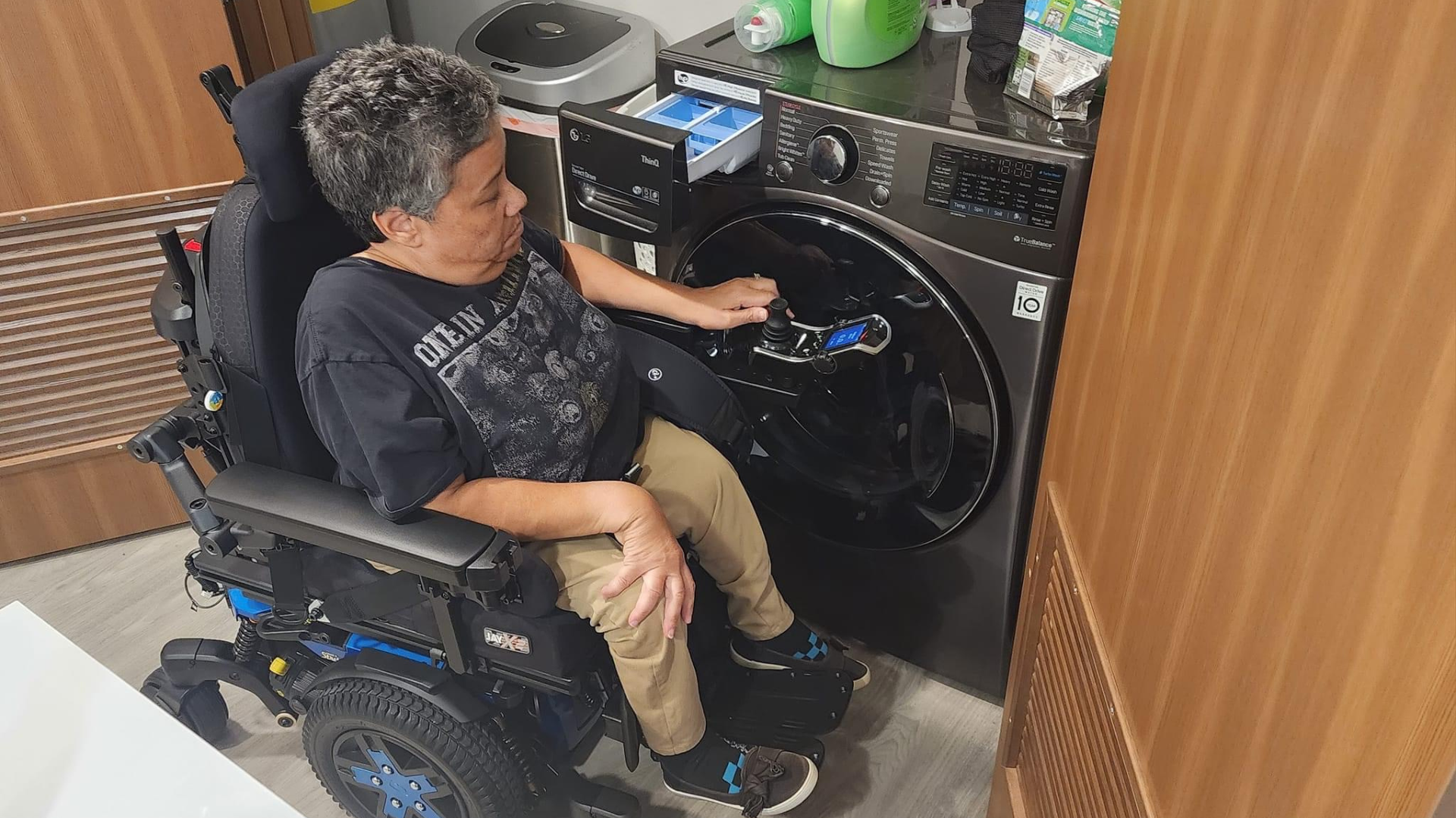 A person in a motorized wheelchair loads laundry into a front-loading washing machine in a compact, accessible room with wooden cabinets and cleaning supplies on top of the washer.