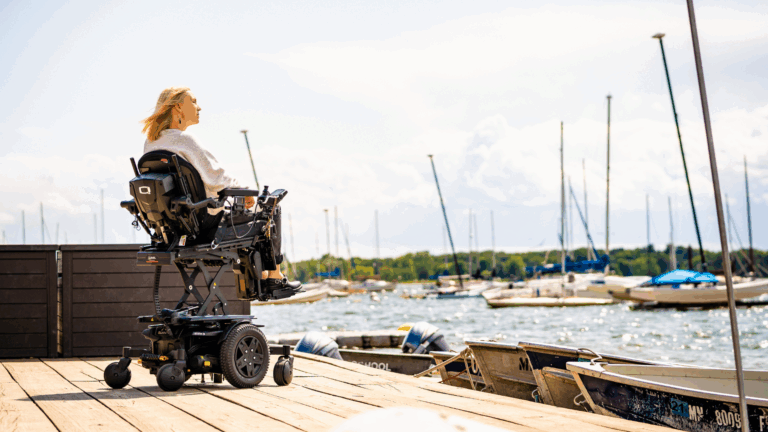 A woman in a powered wheelchair sits on a wooden dock, enjoying accessible activities and looking out over a marina with boats and sailboats under a partly cloudy sky.