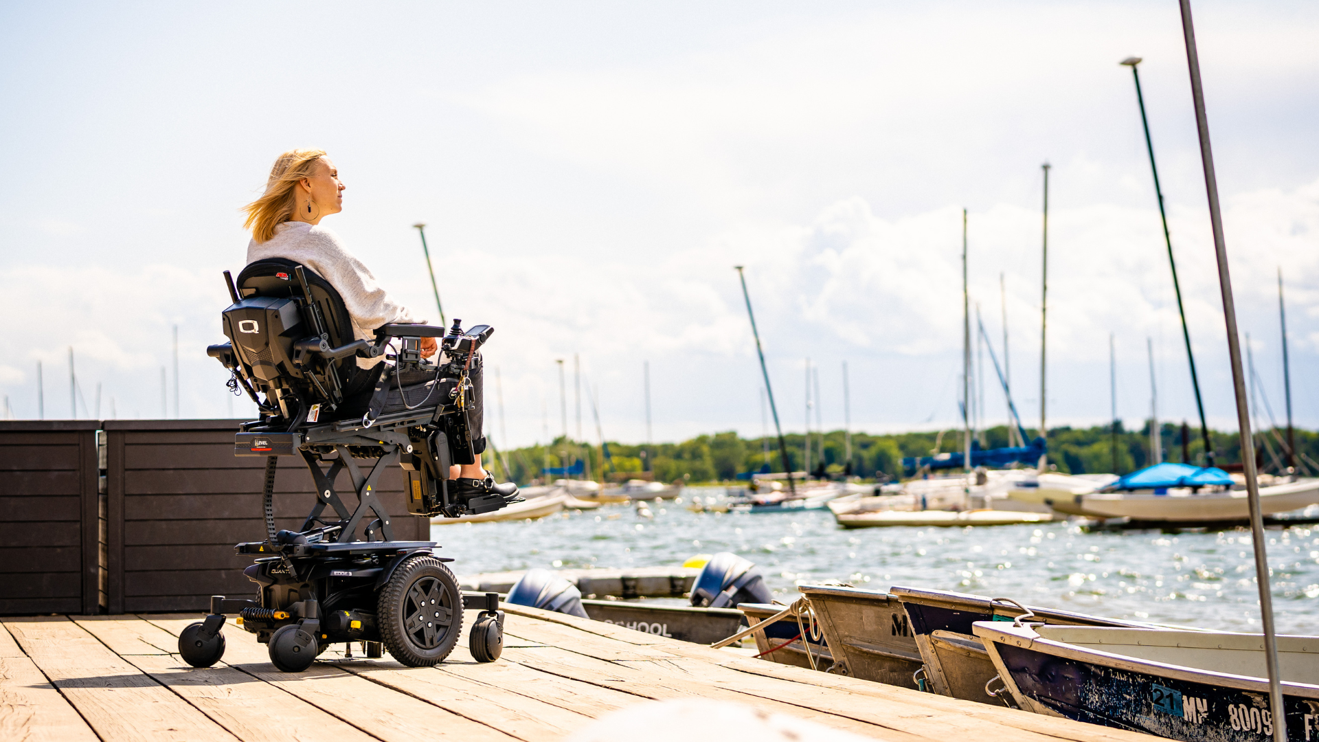 A woman in a powered wheelchair sits on a wooden dock, enjoying accessible activities and looking out over a marina with boats and sailboats under a partly cloudy sky.