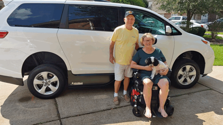 An older man stands beside a woman in a power wheelchair holding a black and white dog, both smiling in front of a white accessible van with adaptive driving features parked in a driveway on a sunny day.