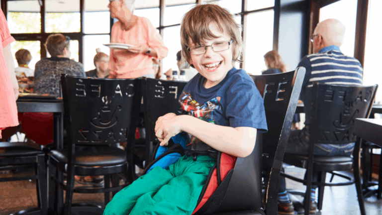A smiling young boy with glasses sits in a high chair at a busy restaurant, wearing a dark blue shirt and green pants. Other people are dining while a woman stands in the background holding plates.