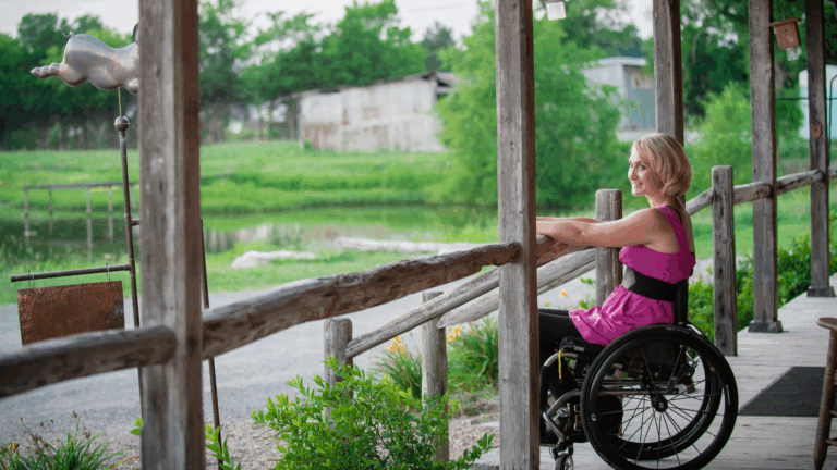 A woman in a wheelchair, dressed in a pink dress, smiles on a wooden porch overlooking a green landscape with trees and a pond. She enjoys the fresh air while keeping up with wheelchair maintenance like clean Velcro for comfort.