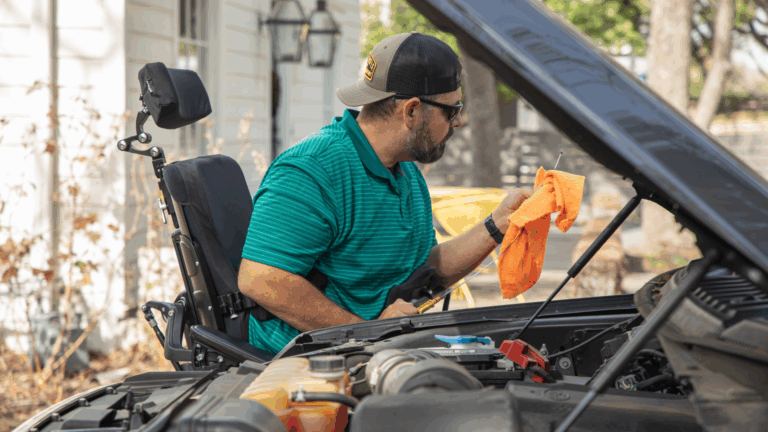 A man in a wheelchair is cleaning the engine of a car with an orange cloth. Wearing a green striped shirt, sunglasses, and a backward cap, he works outdoors in an accessible garage focused on his task.