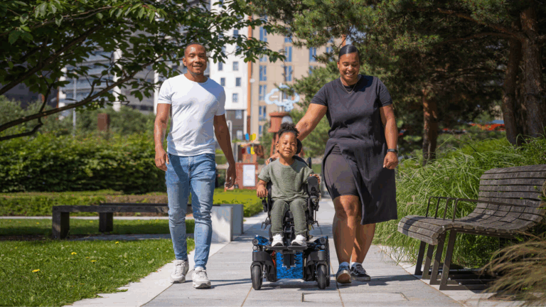A smiling family enjoys a sunny day outdoors; a child with disabilities is pushed by an adult woman, while an adult man walks beside them in a park filled with trees and benches, reflecting the joys of disability parenting.