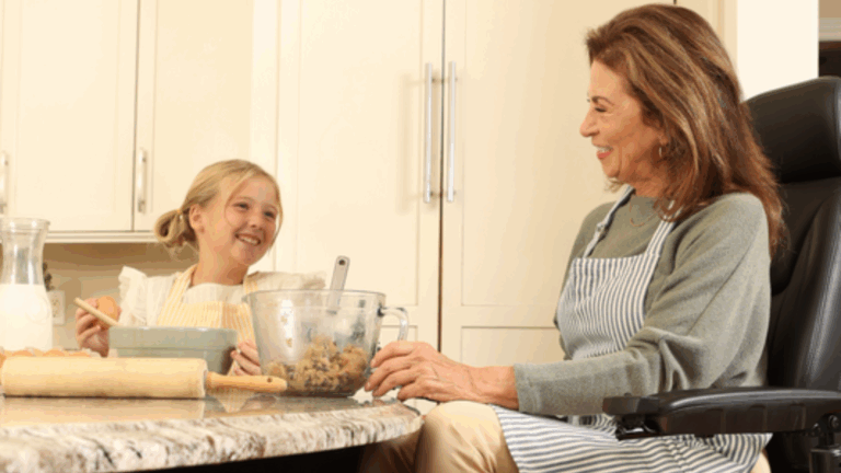A young girl and an older woman wearing aprons smile at each other while baking together in a kitchen. Cookie dough, milk, and baking utensils are on the counter as they chat about taking out the trash after baking.