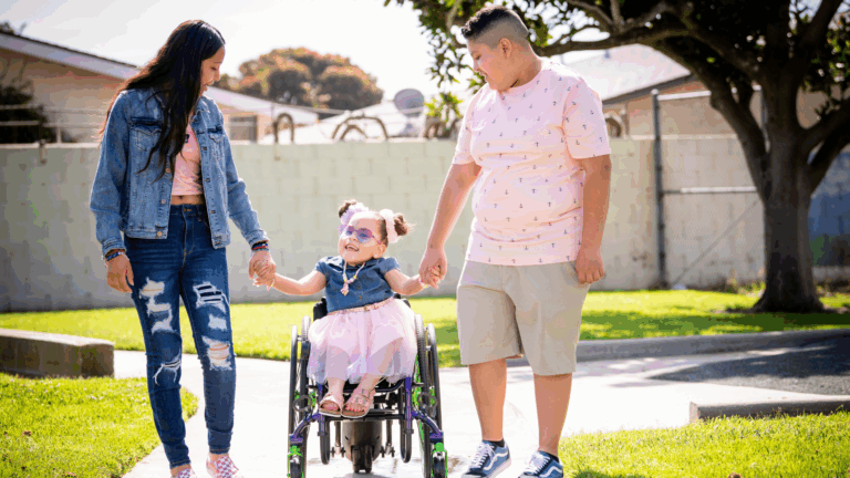 A young girl in a wheelchair smiles while holding hands with a woman and a man outdoors on a sunny day. The happy trio enjoys time together on a sidewalk in a grassy area, highlighting the joys of parenting.
