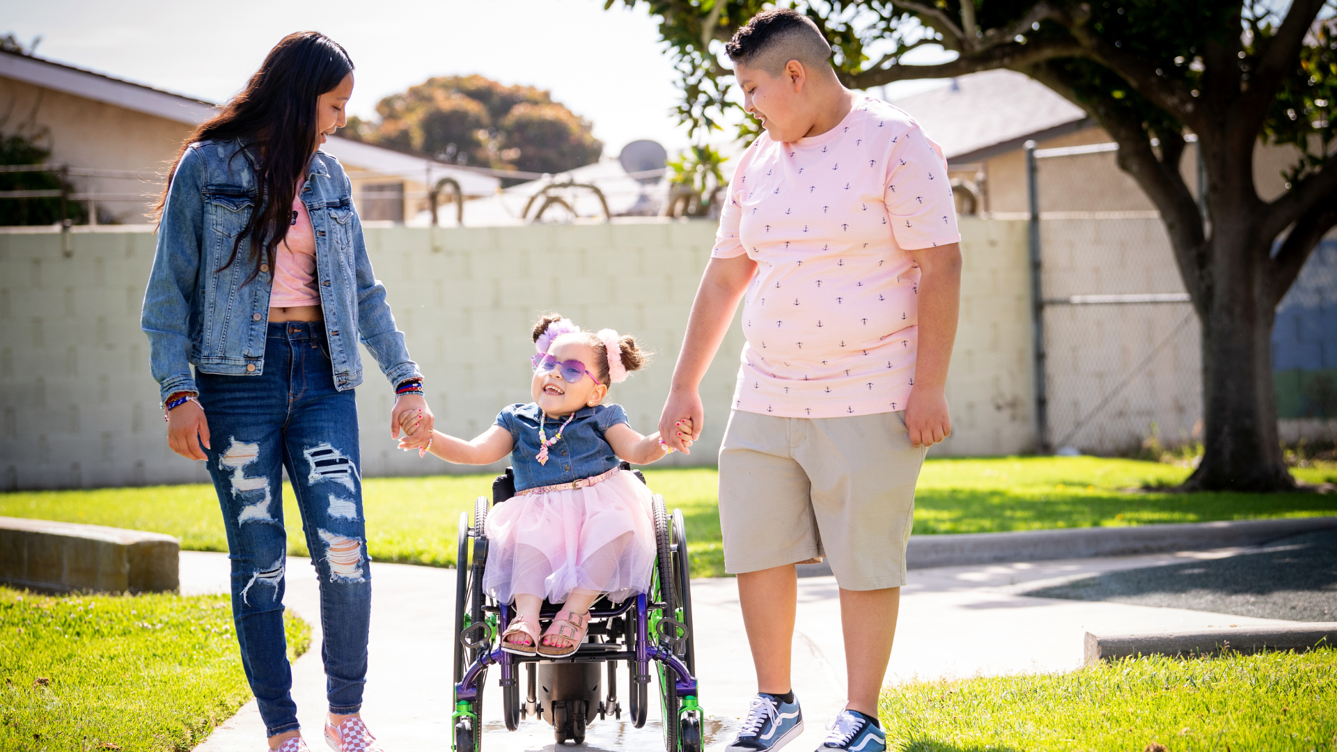 A young girl in a wheelchair smiles while holding hands with a woman and a man outdoors on a sunny day. The happy trio enjoys time together on a sidewalk in a grassy area, highlighting the joys of parenting.