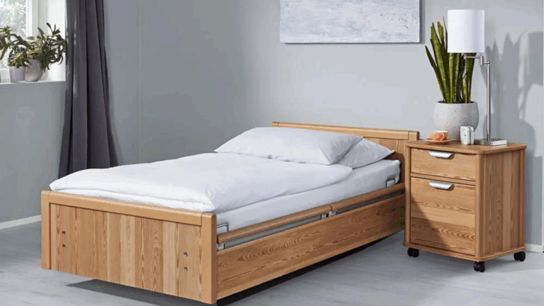 A neatly made single wooden bed with white bedding sits next to a wooden nightstand holding a lamp, potted plant, mug, and two books. There are green plants on the windowsill and a painting on a gray wall, emphasizing sleep importance for disability health.