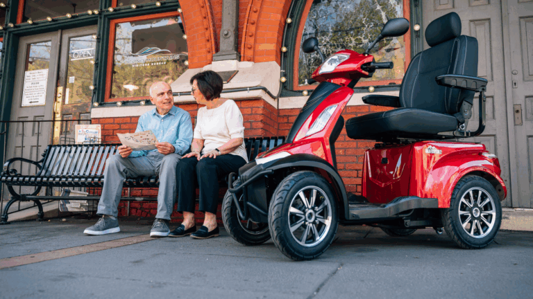 An older man and woman sit on a bench outside a brick building, talking and holding a newspaper. A red mobility scooter, often used for wheelchair travel, is parked in front of them on the sidewalk.