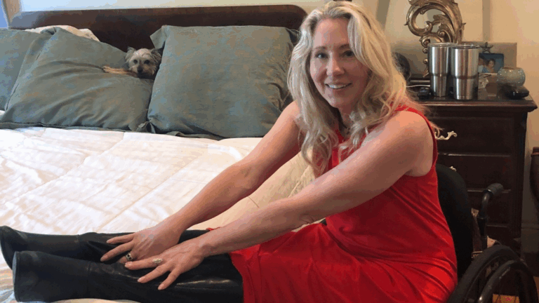 A woman in a red dress sits in a wheelchair beside a bed, smiling at the camera while stretching her arms toward her black boots. A small dog peeks out from between pillows on the bed, illustrating a High Bed Transfer setting.