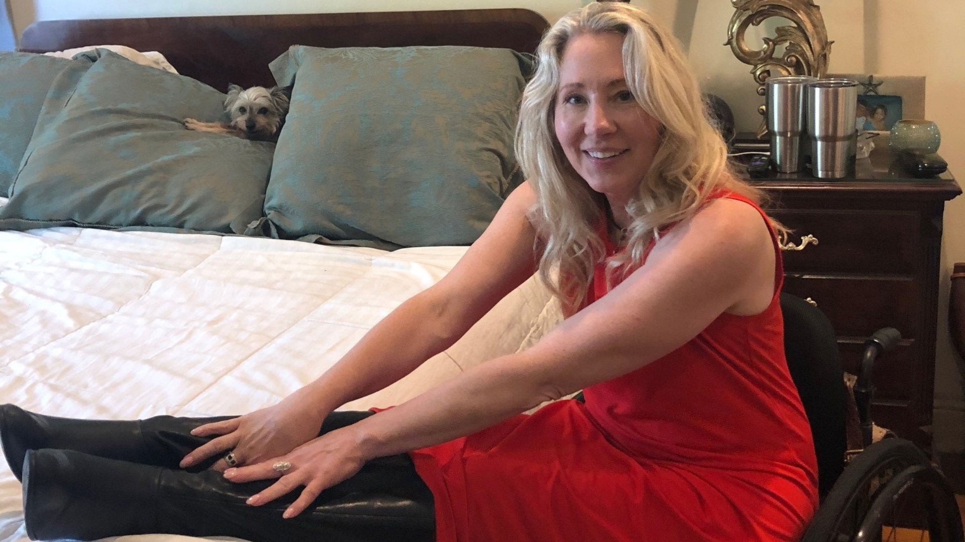 A woman in a red dress sits in a wheelchair beside a bed, smiling at the camera while stretching her arms toward her black boots. A small dog peeks out from between pillows on the bed, illustrating a High Bed Transfer setting.