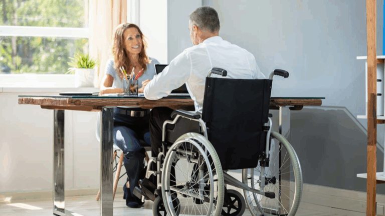 A man using a wheelchair sits at a desk talking to a smiling woman. They are in a bright office with sunlight streaming in and a potted plant on the windowsill, discussing disability employment opportunities.