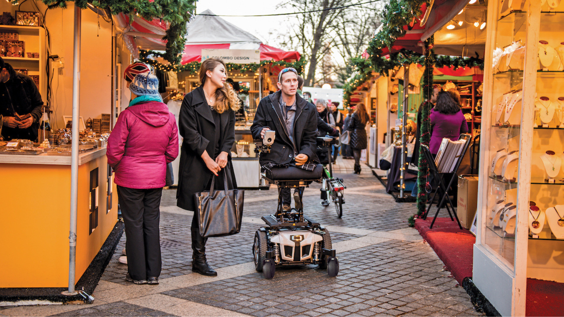 People browse stalls at an outdoor market decorated with garlands during Black Friday shopping. A man in a mobility scooter and a woman walk together, while other shoppers look at jewelry and gifts on display.