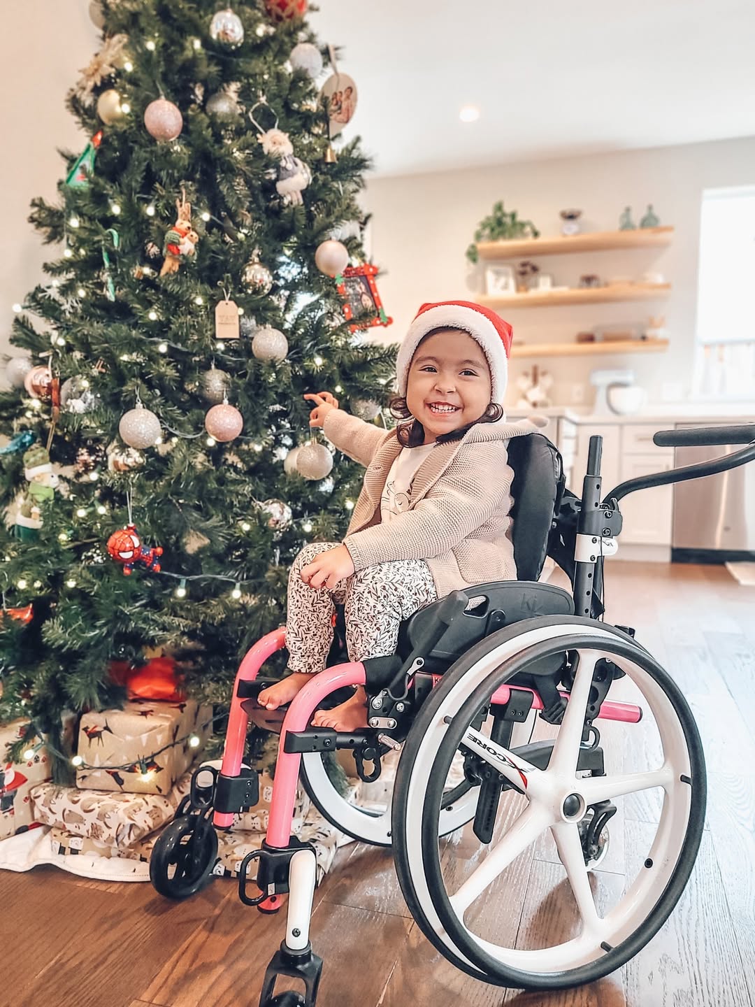 A young child in a wheelchair wearing a Santa hat smiles and points at ornaments on a decorated Christmas tree, with wrapped presents underneath in a cozy, sunlit living room—capturing the joy of simple holiday tips.