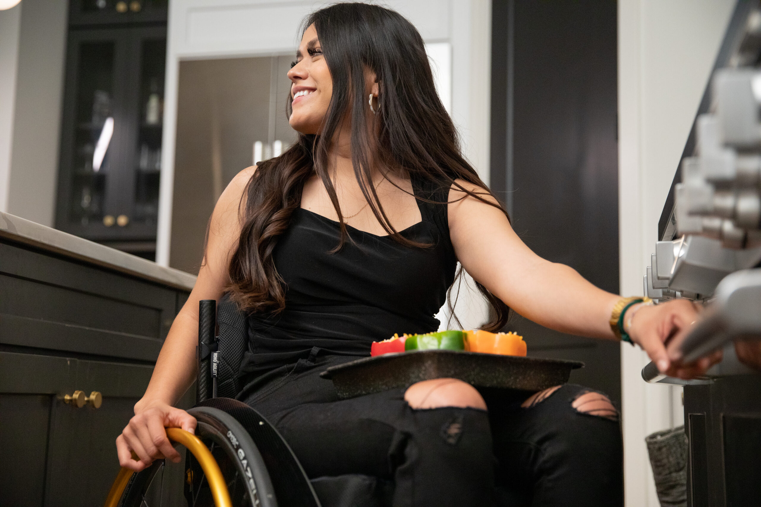 A woman smiles as she sits in a kitchen, reaching towards the oven. She has long dark hair and is holding a tray of colorful vegetables on her lap, highlighting apartment accessibility with thoughtful features for accessible apartments.