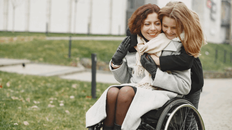 A woman using a wheelchair, wearing a light coat and scarf, smiles as a young girl hugs her from behind. They are outdoors in a park on a cloudy day, enjoying accessible holiday activities together and looking happy and content.