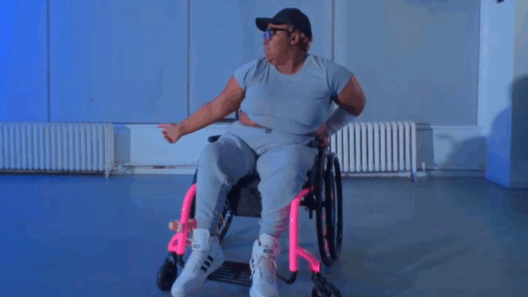 Jamilah Ray, wearing a gray outfit with pink accents, white sneakers, a black cap, and glasses, dances energetically in a spacious room. One arm is raised as she looks to the side, filling the scene with story and movement.