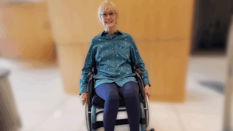 An older woman with short blonde hair, glasses, and a blue button-up shirt sits in a wheelchair, smiling. The softly blurred background with neutral brown and white tones sets the scene for Susans Story.