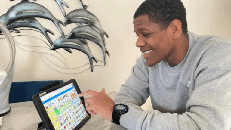 DeVonte smiles while using a communication device with colorful symbol buttons at a table. Behind him, a metallic dolphin sculpture decorates the wall, adding character to his story.