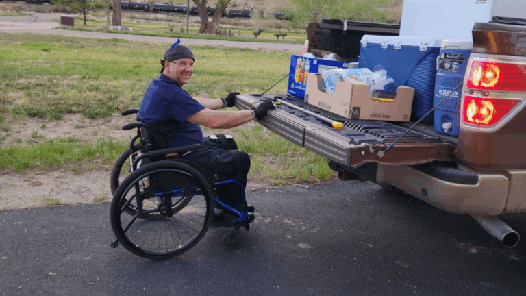 A man in a wheelchair smiles while unloading groceries and supplies from the open tailgate of a pickup truck, parked on a paved area with grass and trees in the background, capturing a moment from Quintens Story.