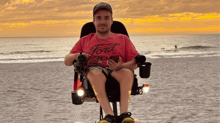 Quintens Story: A man in a power wheelchair sits on a sandy beach at sunset, wearing a red Ford T-shirt, cap, and shorts, holding a smartphone, with the ocean and a yellow-orange sky behind him.