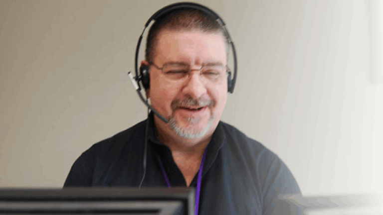 Gregs Story: A man wearing glasses and a headset smiles while working at a computer, suggesting he is providing customer service or technical support.