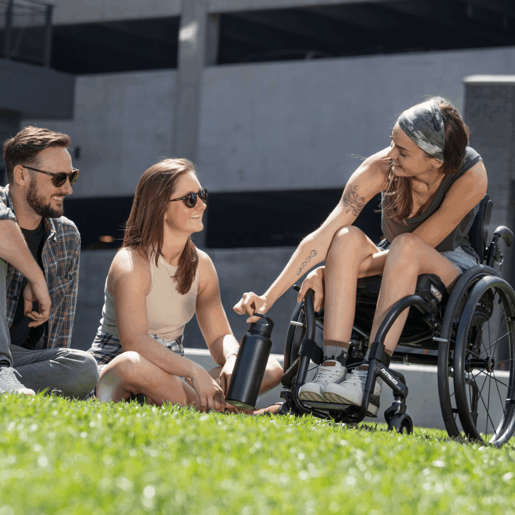 Three young adults enjoying a sunny day outdoors. A woman in a wheelchair smiles while handing a water bottle to a seated woman, with a man beside them. All are relaxed and wearing casual summer clothes.