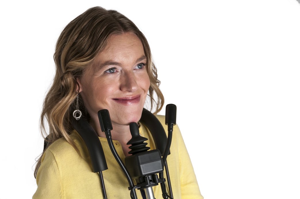 A woman with wavy blonde hair, wearing a yellow sweater, smiles while using a wheelchair equipped with Alternative Access Controls such as a mounted joystick and headrest against a white background.