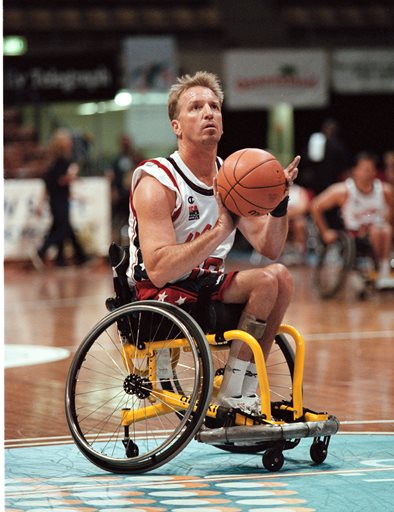 A male adaptive athlete prepares to take a basketball shot during a game on an indoor court. He wears a sports jersey and shorts, with other players visible in the background.