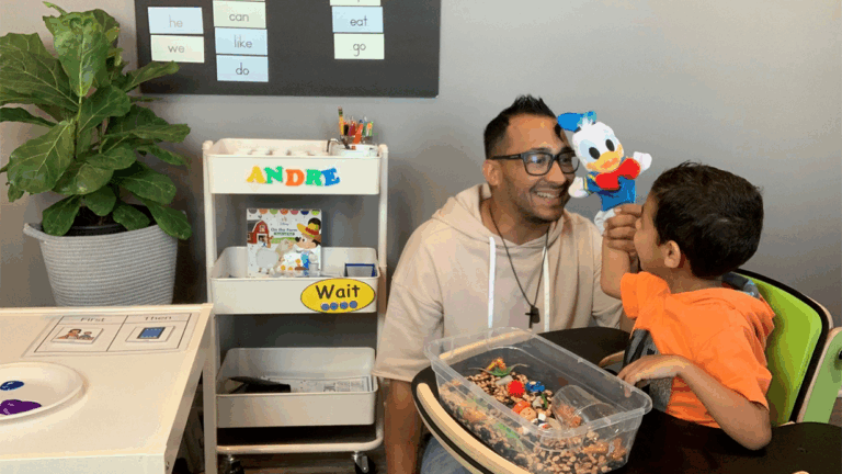 An adult and a child sit in a classroom. The adult smiles as the child holds up a Donald Duck puppet, sharing a moment from Andres Story. Nearby are a sensory bin, activity table, Andre-labeled cart, plant, and educational posters.