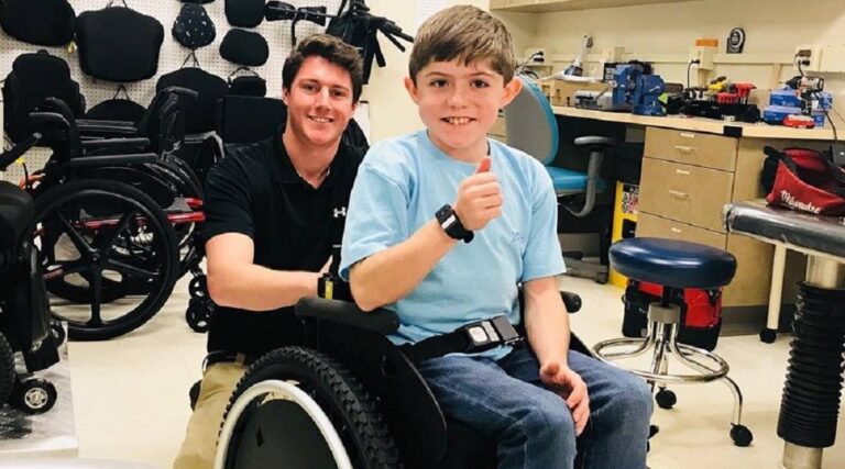 A young boy in a wheelchair gives a thumbs up, smiling, while an adult man kneels behind him in a workshop filled with wheelchairs and equipment—capturing the spirit of Andrews Story.