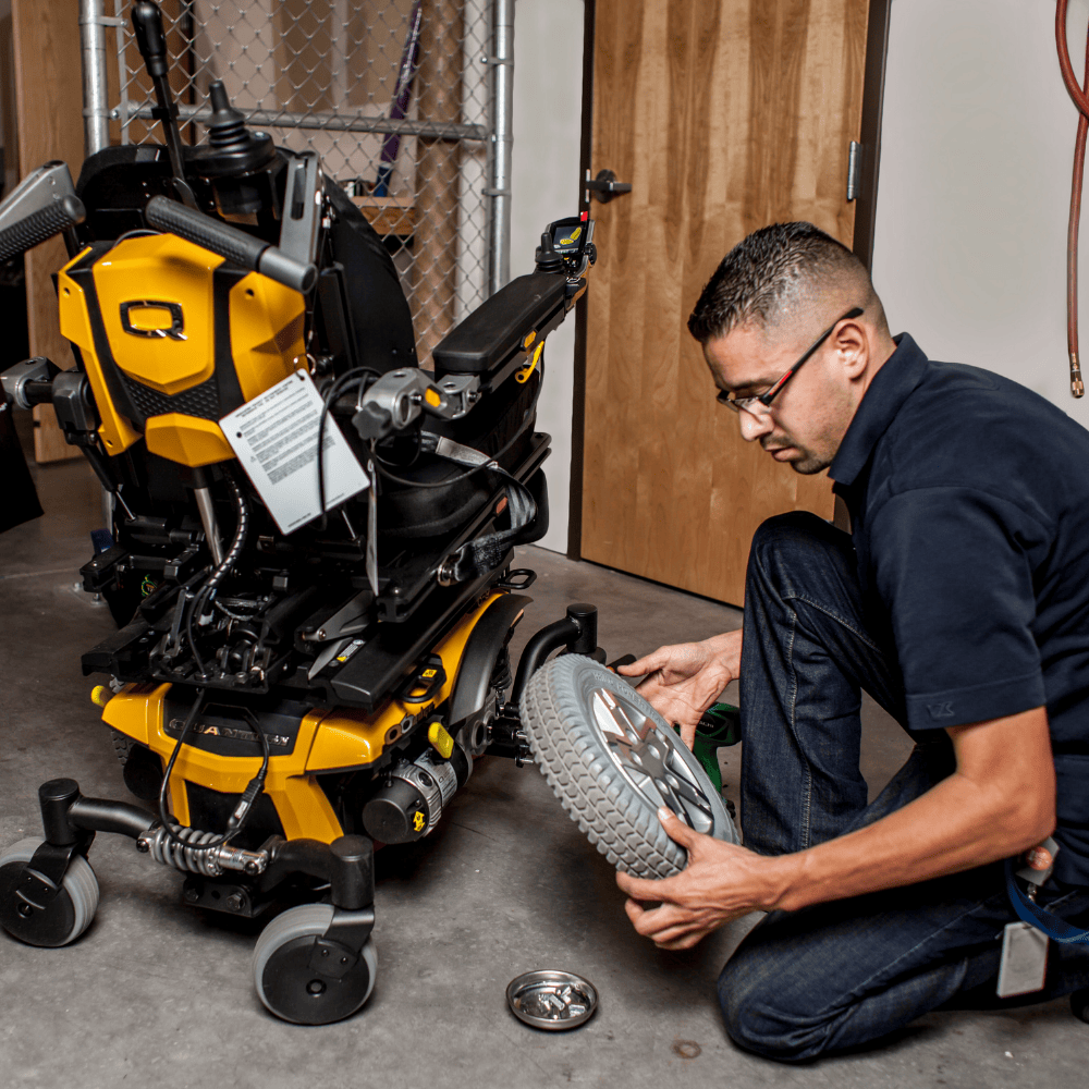 A technician repairs a yellow power wheelchair in a workshop, carefully inspecting a detached rear wheel. Tools and parts are on the floor nearby, and the setting includes a wooden door and metal chain-link partition.