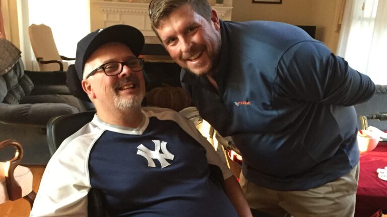 Two men smiling indoors. One man, featured in Brians Story, wears glasses, a Yankees shirt, and a cap. The other stands beside him in a blue zip-up jacket. They share a joyful moment together in a cozy living room.