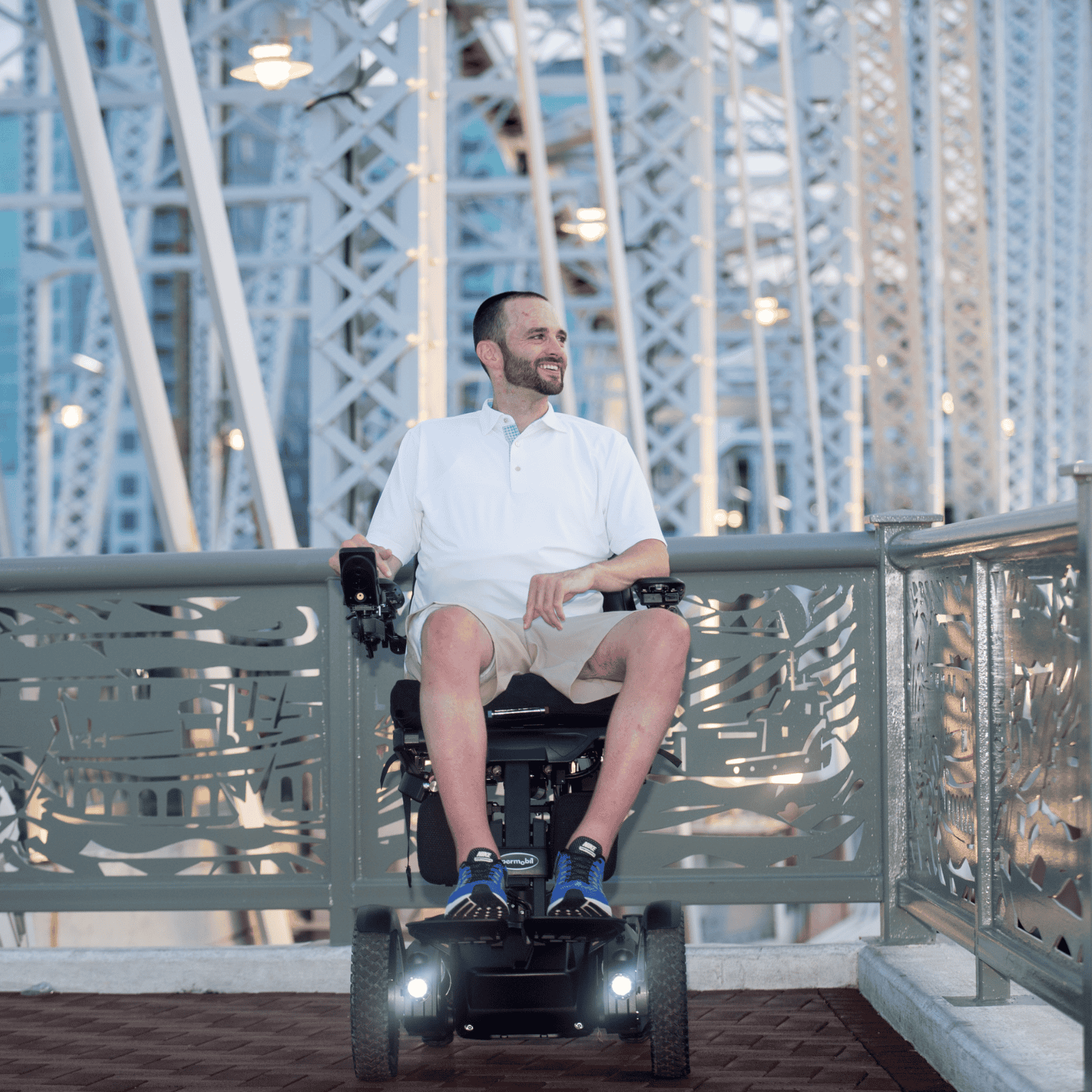 A smiling man in a power wheelchair sits on a bridge at dusk, surrounded by a framework of metal beams and soft lights. He wears a white polo shirt and khaki shorts, enjoying the evening view.