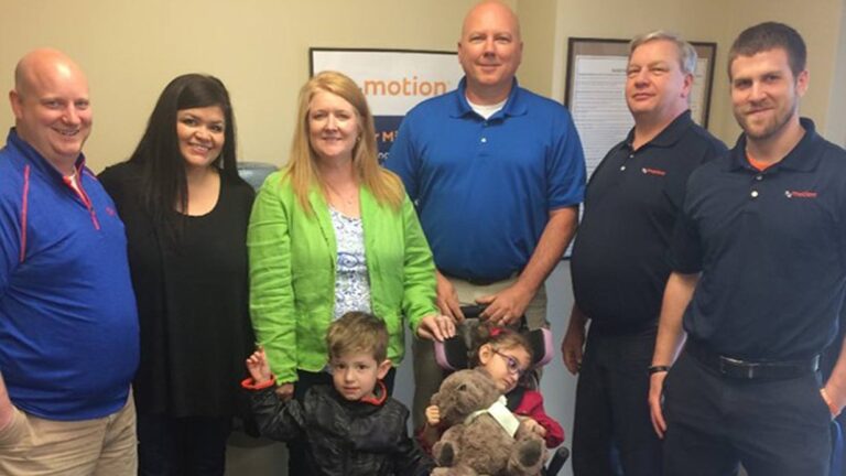 A group of five adults stands behind two young children; one child is holding a stuffed animal. Everyone is smiling and posing together indoors, in front of a wall with a framed Charleys Story poster.