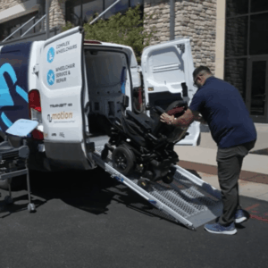 A technician loads a power wheelchair into the back of a white NuMotion service van using a metal ramp. The van is parked outside a building and labeled for complex wheelchair service and repair.