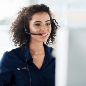 A smiling customer service representative wearing a headset and navy NuMotion shirt assists a client at a desk. She is looking at a computer monitor in a brightly lit office.
