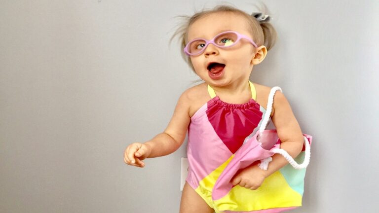 A young child wearing pink glasses and a colorful pink, yellow, and aqua swimsuit smiles excitedly while holding a matching bag, standing against a plain light gray background—celebrating Emerys Story.