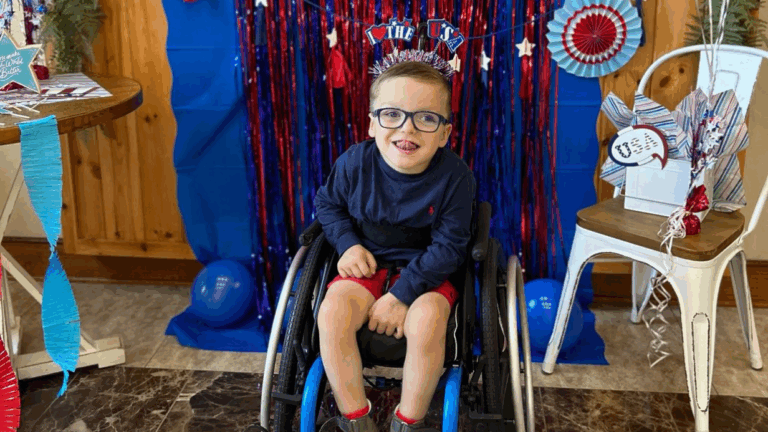 Logans Story: A young boy in a wheelchair, smiling and wearing glasses, sits in front of festive red, white, and blue decorations with streamers and balloons at a patriotic-themed celebration.