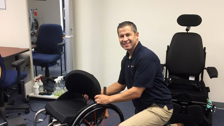 A man kneels next to a wheelchair in an office, adjusting its seat. Smiling at the camera, he shares Marks Story. An empty office chair and a larger medical chair are in the background, with cleaning supplies on a table nearby.