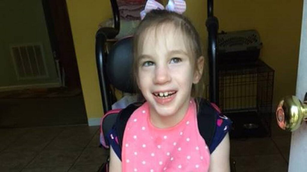 A young girl with light brown hair in a pink bow smiles while sitting in a wheelchair, wearing a pink bib with white polka dots. Indoors, near a yellow wall and door, she’s ready for another adventure in the story of Mikah & Izzy.